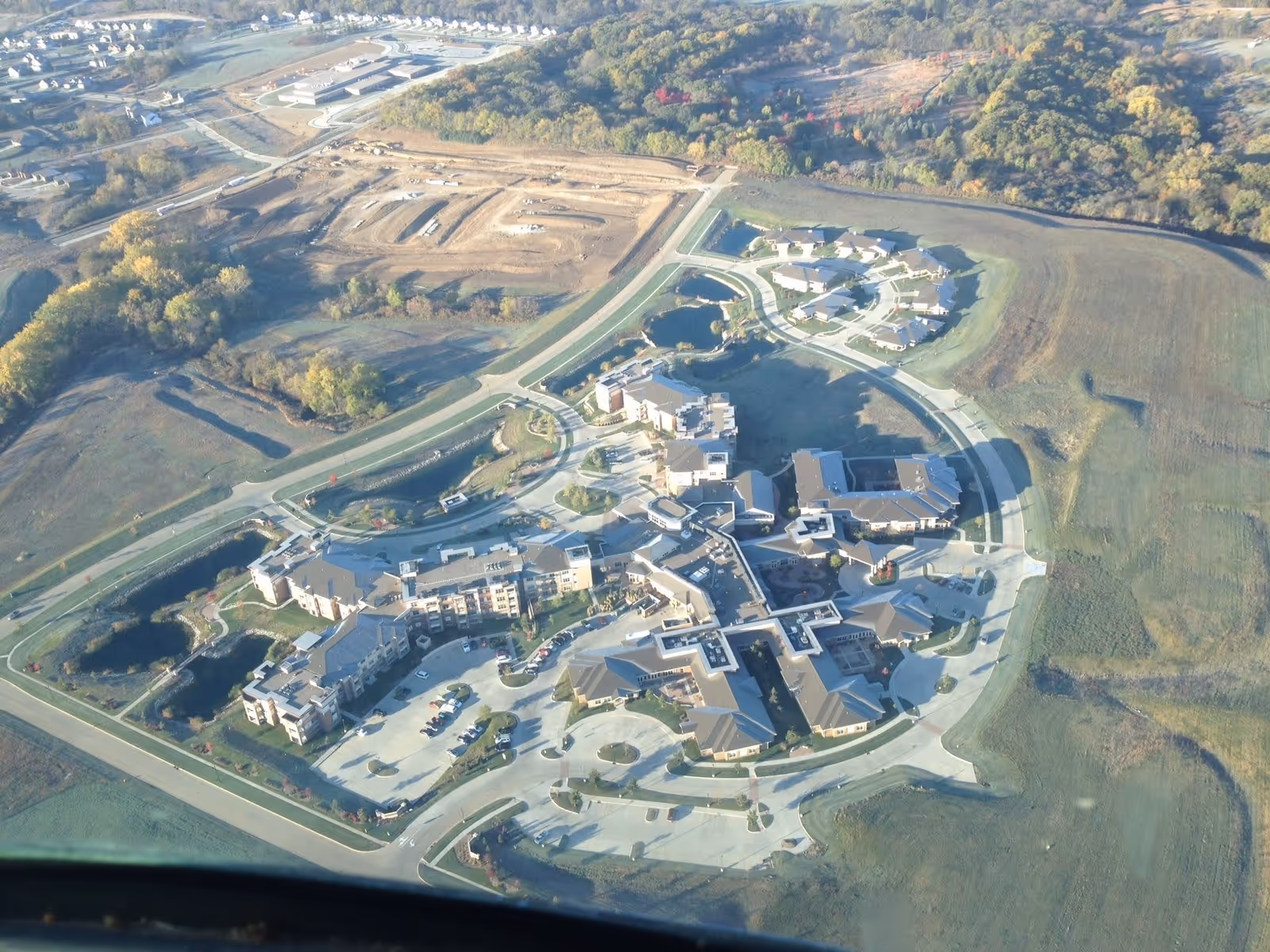 Aerial view of Edgewater, a WesleyLife Community for Healthy Living, showing a large senior living facility complex surrounded by roads, parking areas, green fields, and some wooded areas in the background.