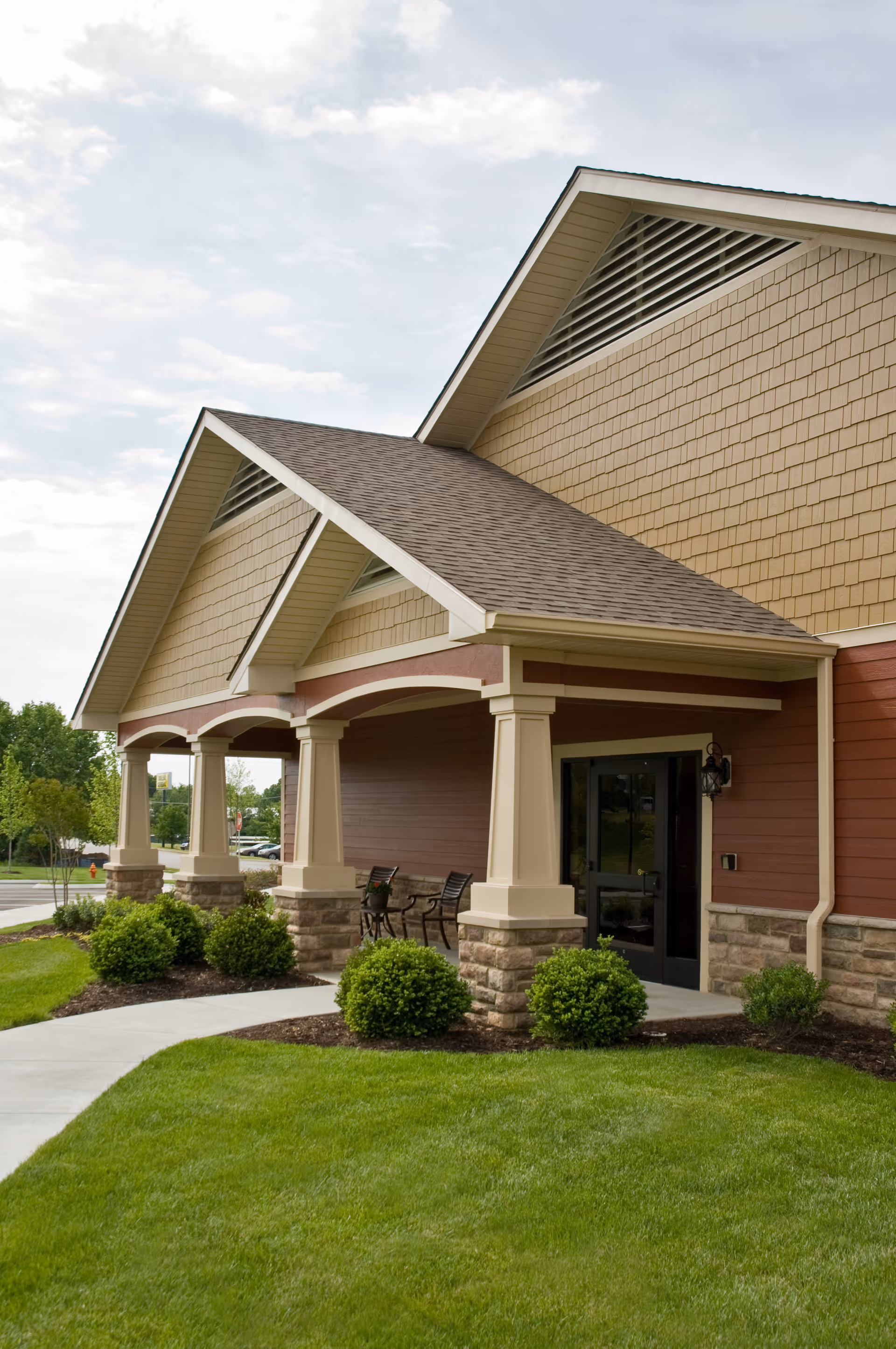 Covered front entrance of a building with columns, outdoor seating, and manicured lawn and shrubs.