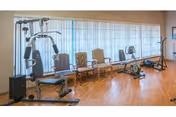 A senior fitness room with a weight machine, stationary bike, and chairs along a windowed wall.