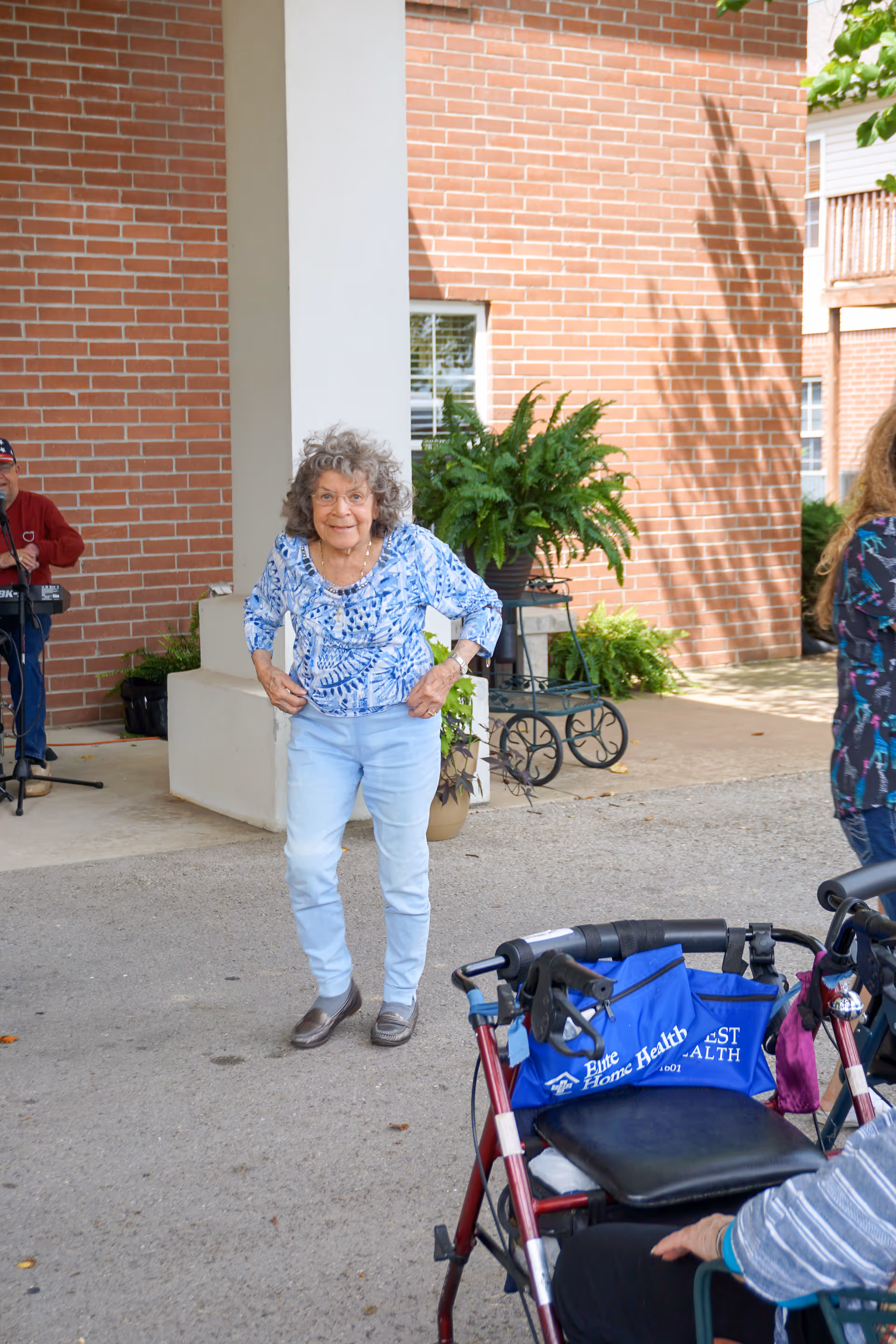 An elderly woman with curly gray hair wearing a blue patterned blouse and light blue pants stands outdoors near a brick building with a white column. In the foreground, there is a red walker with a blue bag attached. Another person is partially visible sitting near the walker. There are plants and a man playing a keyboard in the background.