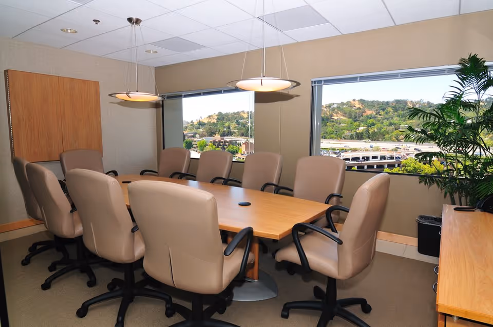 Boardroom-style conference room with a long wooden table, beige chairs, and large windows.