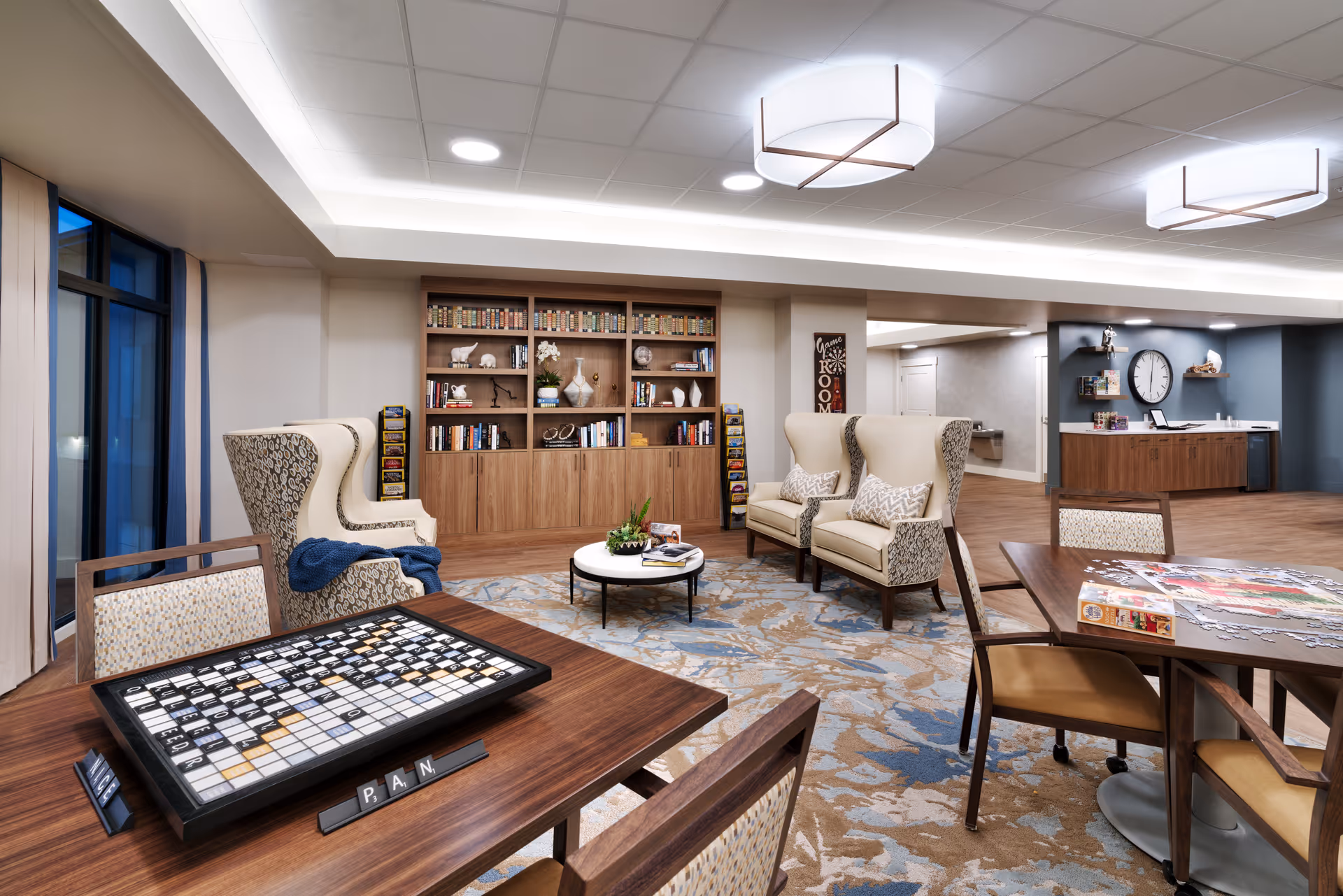 A cozy senior living common area with two tables featuring board games and puzzles, two beige wingback chairs with patterned sides, a large bookshelf filled with books and decorative items, and a carpet with a floral pattern. The room has large windows with curtains, modern ceiling lights, and a wall clock above a counter in the background.
