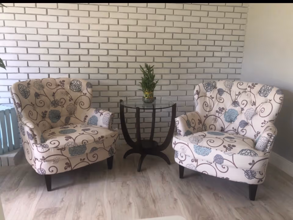 Two patterned armchairs surround a glass table with a plant against a brick wall.