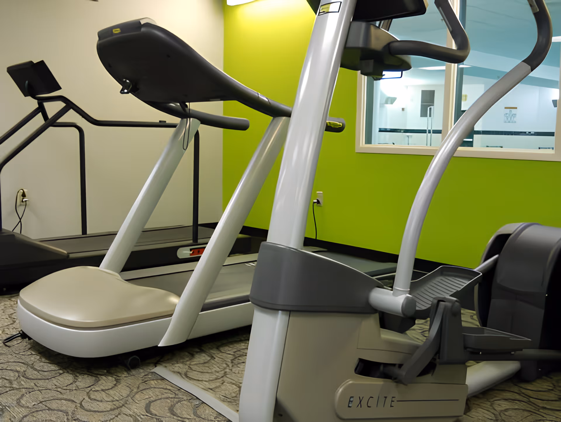 Treadmill and elliptical machines in a small fitness room with a bright green accent wall and interior window.