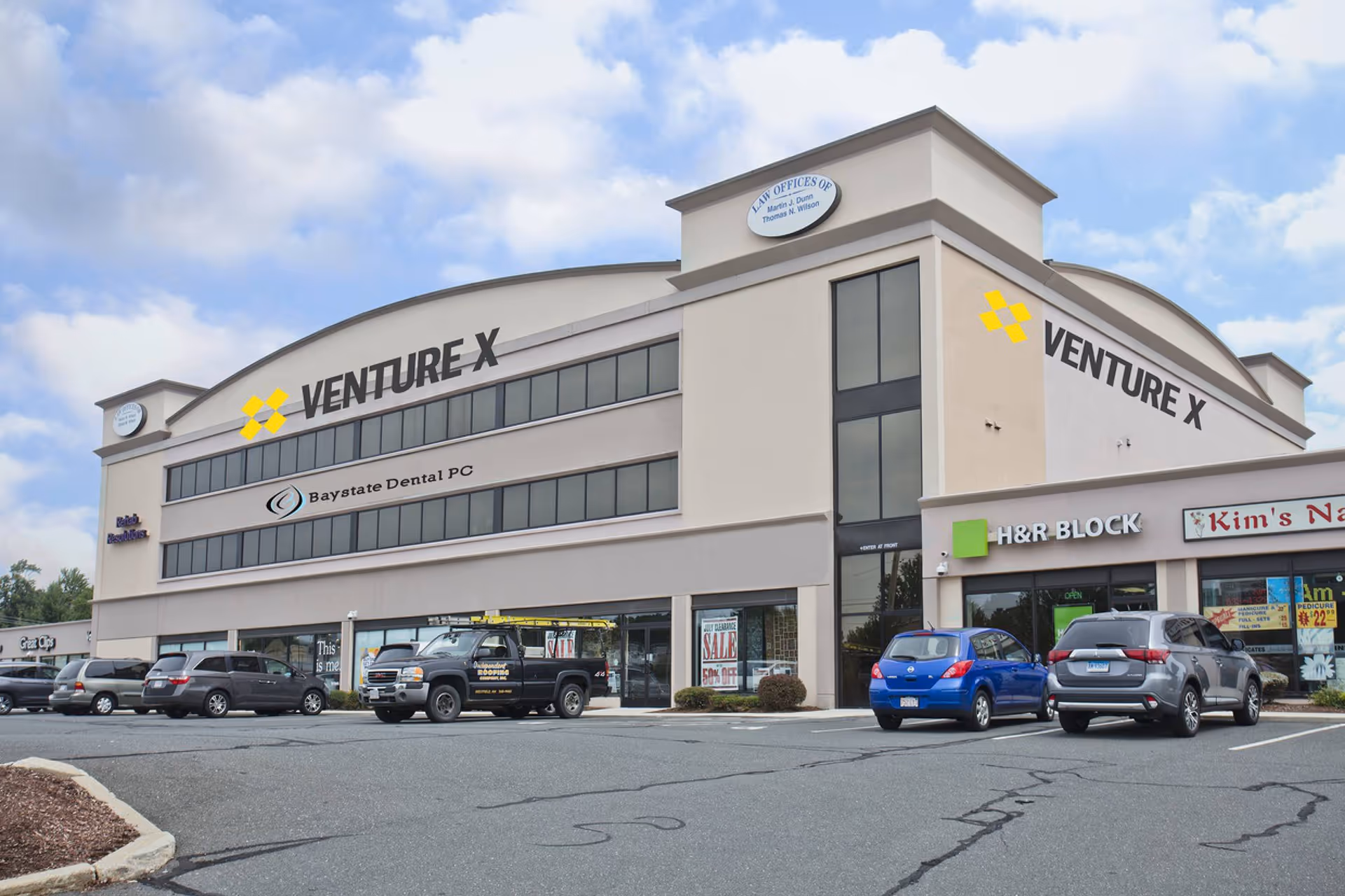 A two-story shopping plaza with multiple storefronts and parked cars in the lot.