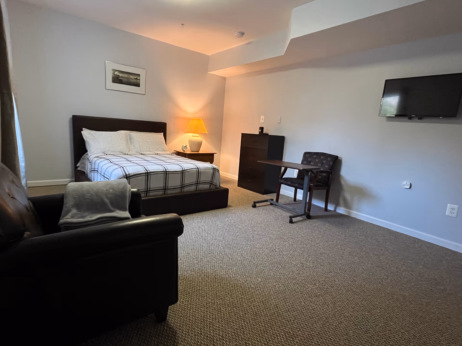 A bedroom with a bed covered in a checkered blanket, a nightstand with a lamp, a dresser, a small table with a chair, a wall-mounted TV, and a cushioned armchair with a folded blanket on it. The walls are light gray and there is a framed picture above the bed.
