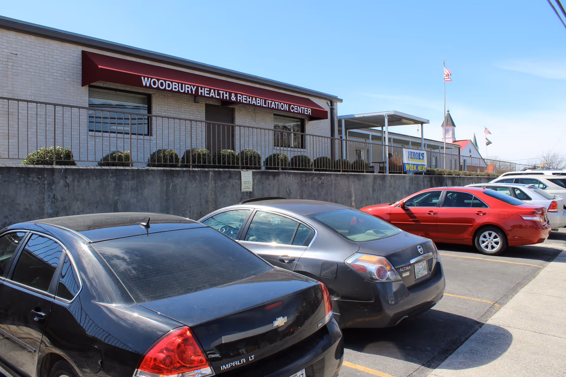 Exterior view of Woodbury Health & Rehabilitation Center showing a single-story building with a red awning above the entrance. Several cars are parked in the parking lot in front of the building. An American flag is visible in the background against a clear blue sky.