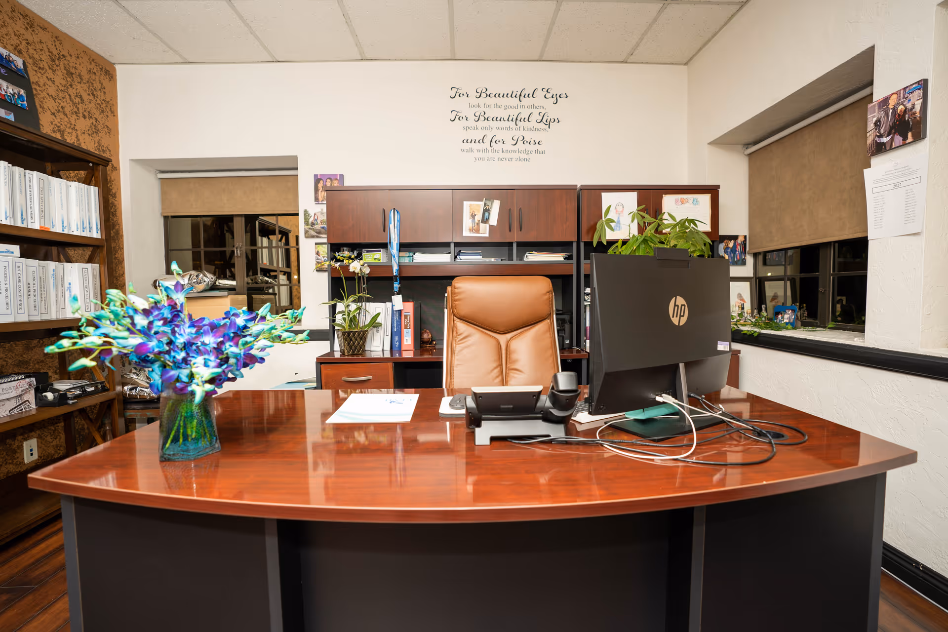 A polished wooden reception desk with computer, chair, plants, and filing shelves in an office.