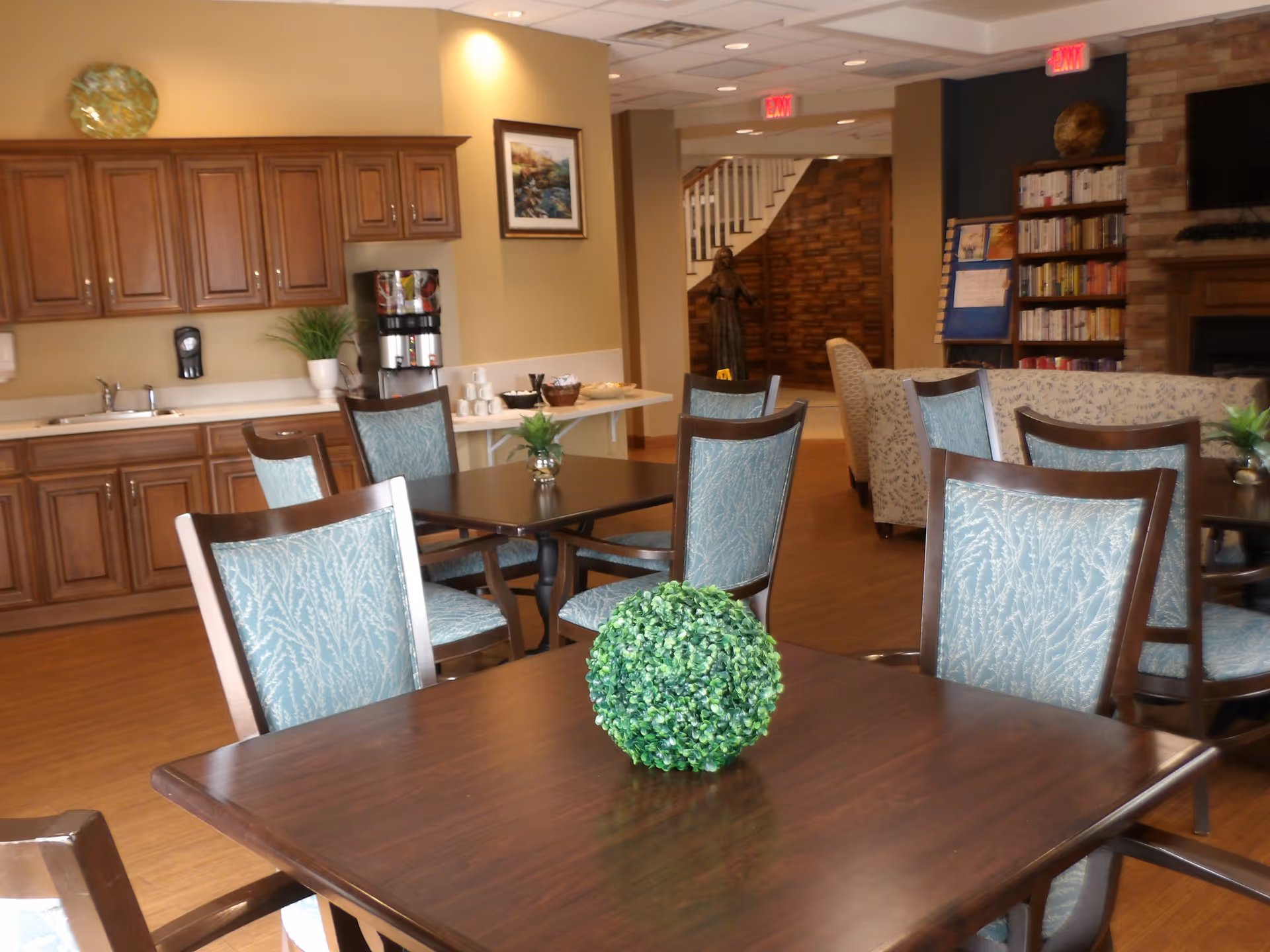 Communal dining and lounge area with wooden tables, teal-upholstered chairs, a kitchenette and bookshelves in the background.