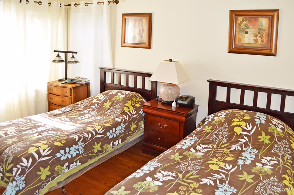 A bright bedroom with two single beds featuring brown bedspreads with green, blue, and white floral patterns. Between the beds is a wooden nightstand with a lamp, a clock, and a small figurine. There is a wooden dresser with books and a lamp near a window with white curtains. Two framed pictures hang on the wall above the beds.