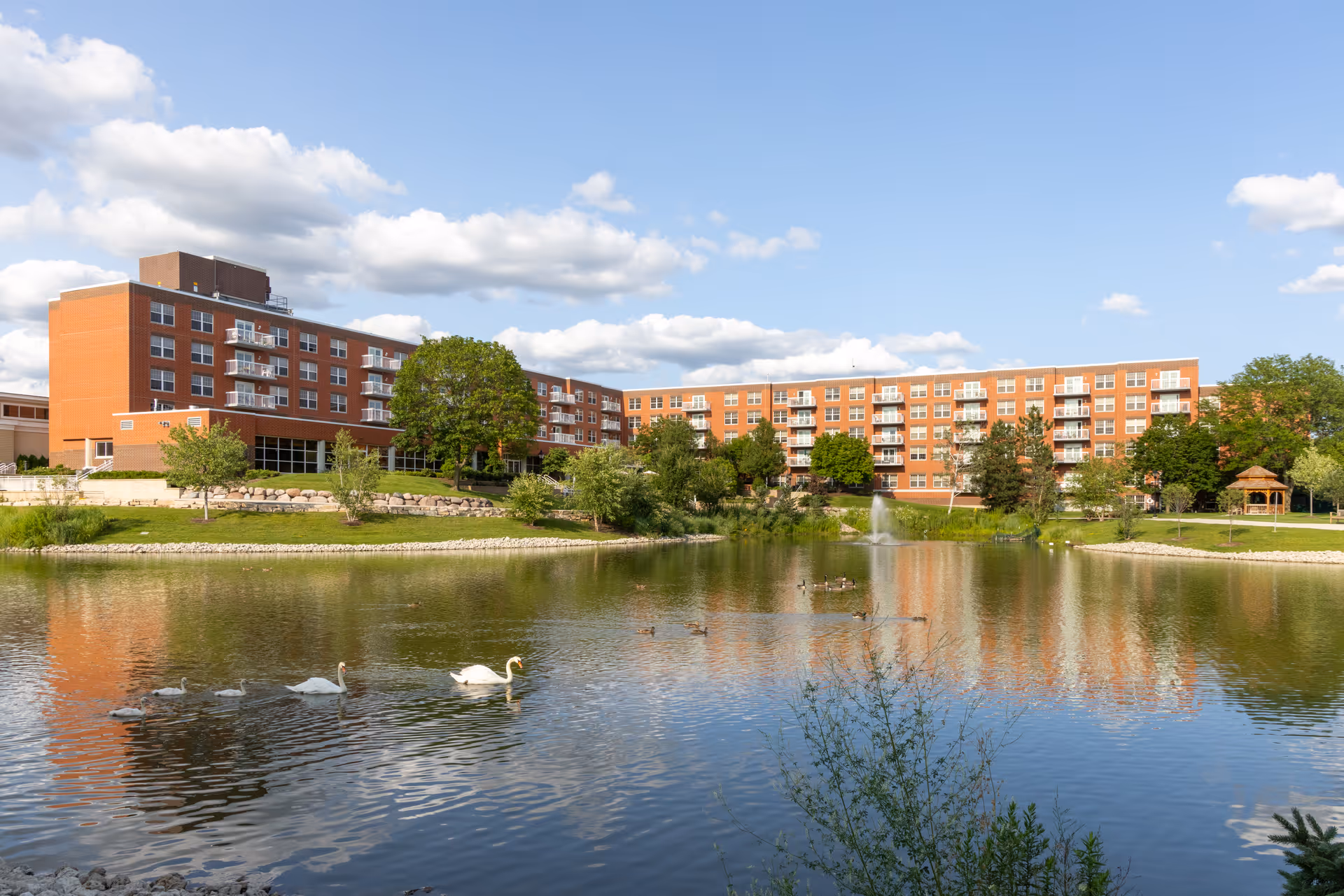 Large multi-story brick senior living building reflected in a pond with swans and a fountain under a blue sky.