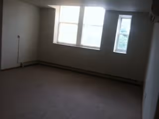 Empty carpeted room with two windows, baseboard heating, and plain white walls.