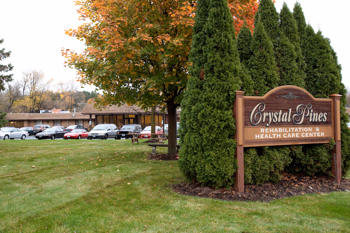 Exterior view of Crystal Pines Rehabilitation & Health Care Center with a wooden sign in front surrounded by tall evergreen trees and a large tree with autumn-colored leaves. Several cars are parked in front of the building.