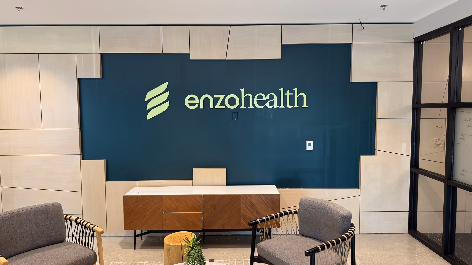 Reception area with teal Enzo Health logo wall, wood paneling, and gray seating.