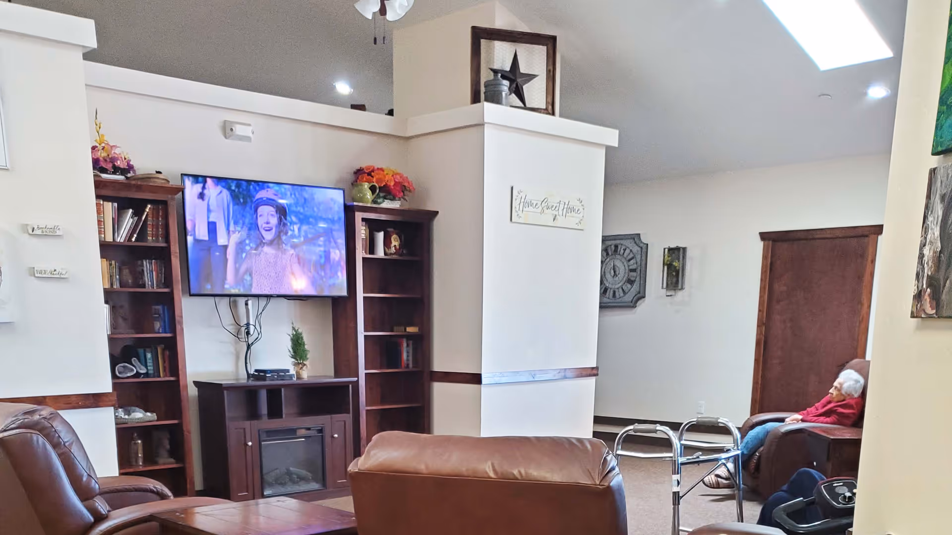 A cozy living room area in a senior living facility with brown leather chairs arranged around a wooden coffee table. A flat-screen TV is mounted on the wall above a wooden cabinet with shelves on either side filled with books and decorative items. A walker is positioned near one of the chairs, and an elderly person is seated in a recliner chair near a wooden door. The room has light-colored walls, a high ceiling with recessed lighting, and a sign on the wall that reads 'Home Sweet Home'.
