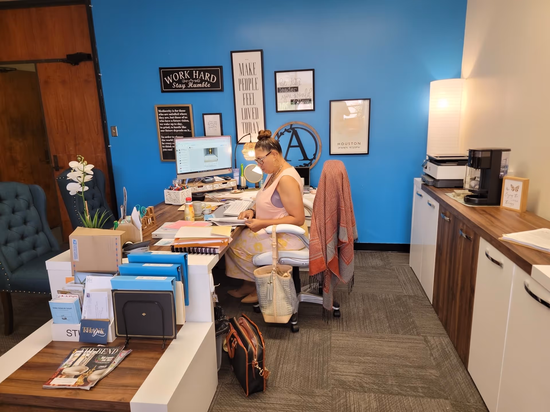 An office desk area with a woman working at a computer against a blue wall.