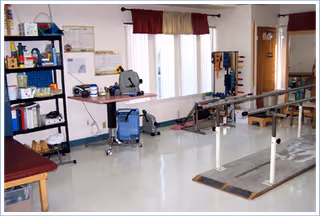Rehabilitation room with parallel walking bars, exercise equipment, shelving, and a treatment table near windows.