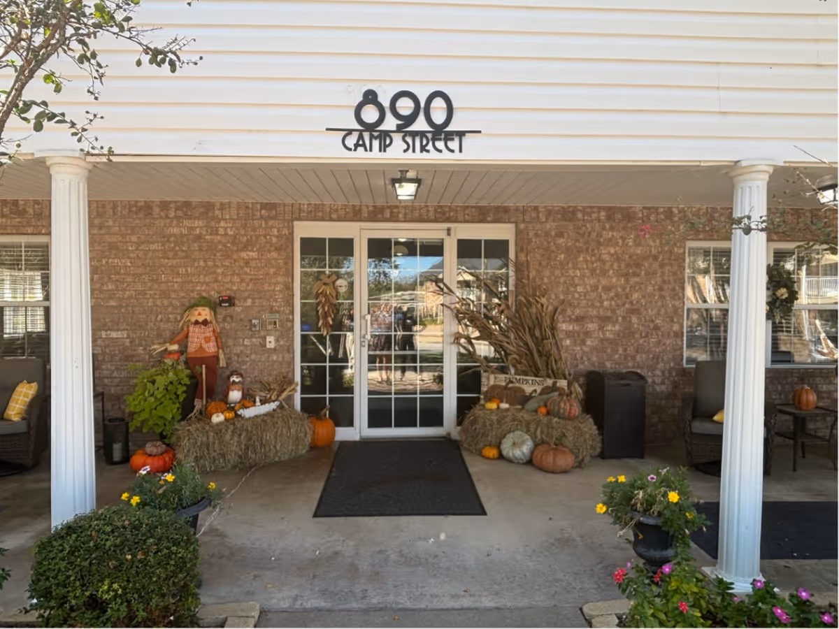 Entrance of a building with the address 890 Camp Street displayed above the door. The entrance is decorated with autumn-themed items including hay bales, pumpkins, corn stalks, and scarecrows. There are two white columns supporting the roof over the entrance, and seating areas with chairs and small tables on either side. Potted plants with flowers are placed near the entrance.
