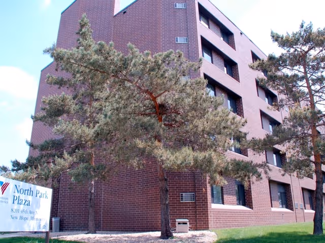 Brick senior living building with large trees and a North Park Plaza sign in front.