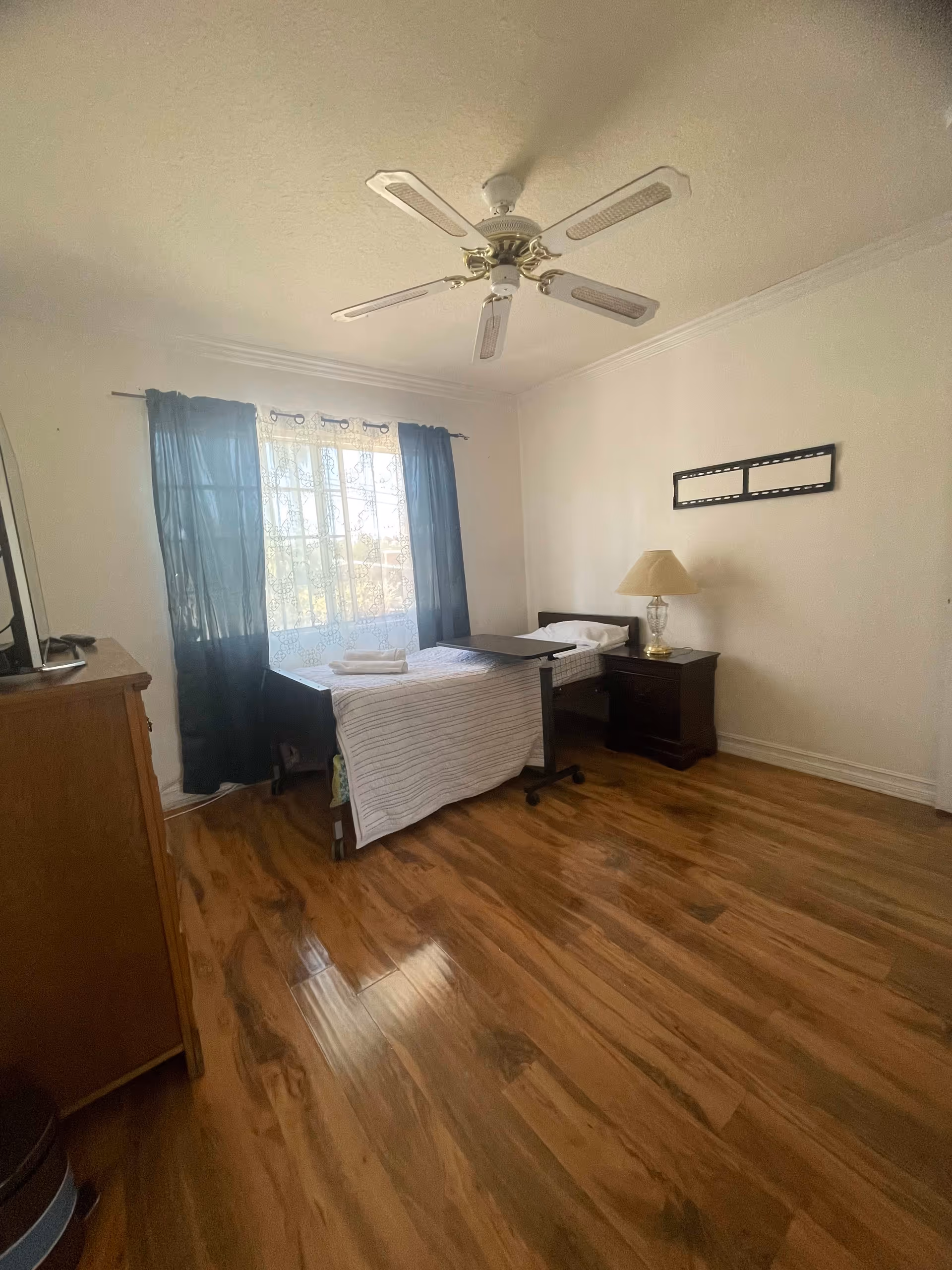 A simple bedroom with a single bed covered with a striped blanket, a small bedside table with a lamp, a ceiling fan, a window with sheer and dark curtains, and a wooden dresser with a TV on top. The floor is wooden and the walls are plain white.