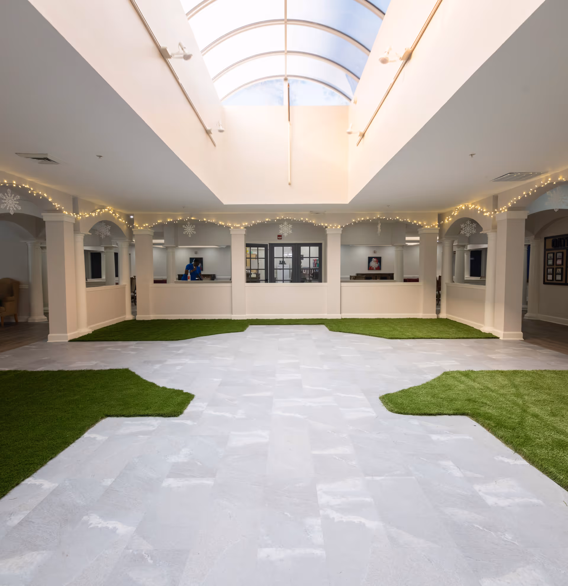 Interior view of a spacious common area with a large skylight ceiling. The floor is a combination of light-colored tiles and patches of green artificial grass. The walls have white columns and arches decorated with string lights and snowflake ornaments. There is a reception or office area with windows and a person visible inside.
