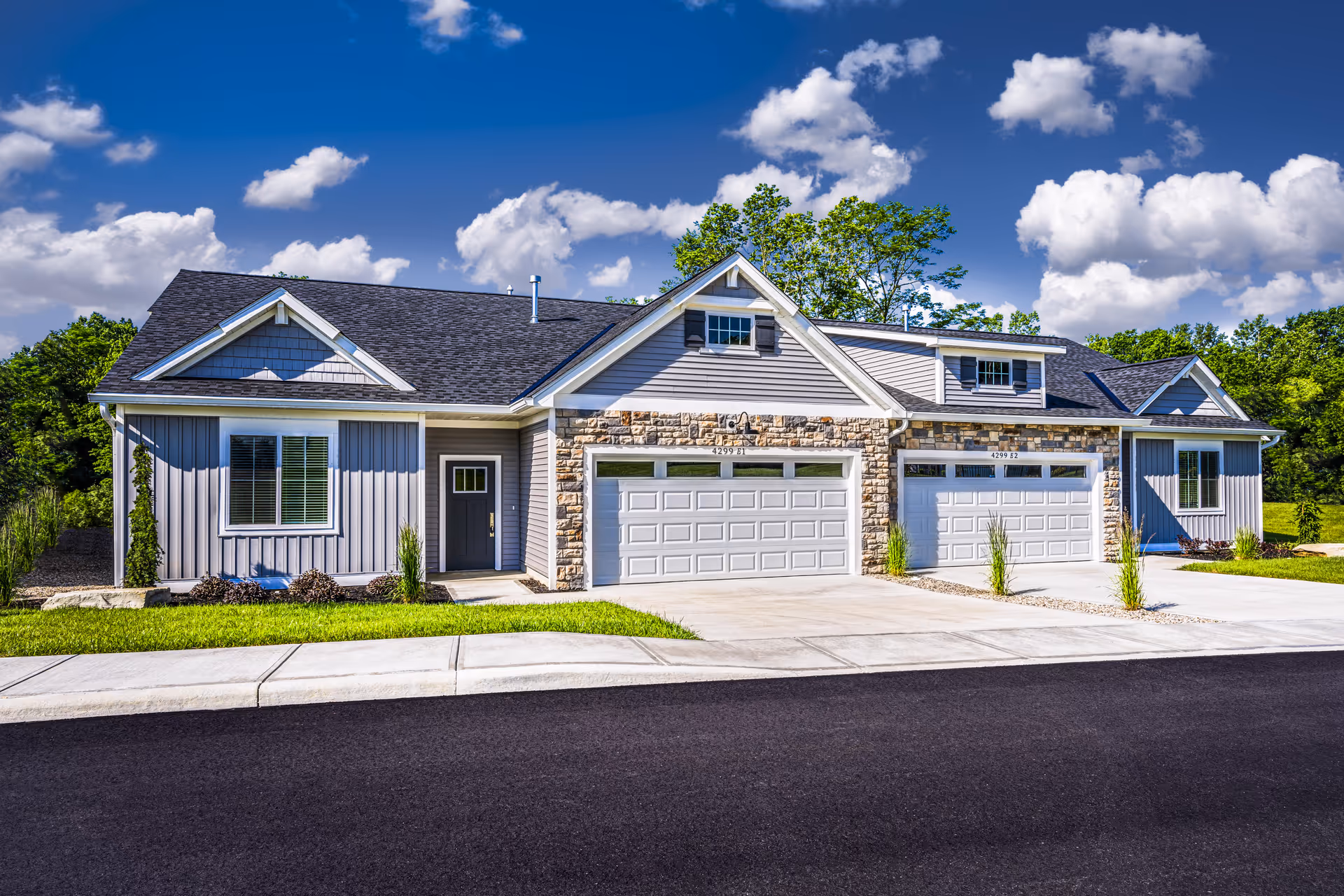 Exterior view of a modern residential building with two attached garages, light gray siding, stone accents, and a dark shingled roof under a bright blue sky with scattered clouds. The building is surrounded by green grass and trees in the background.