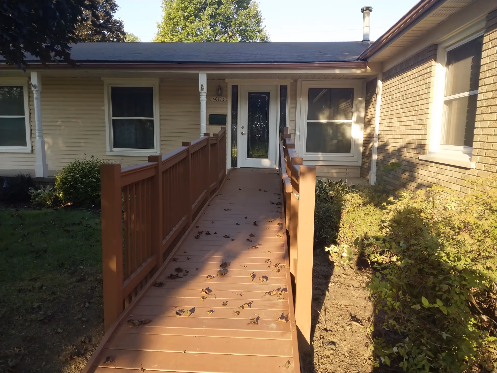 A wooden ramp with handrails and scattered leaves leading to the front entrance of a single-story house with beige siding and brick.