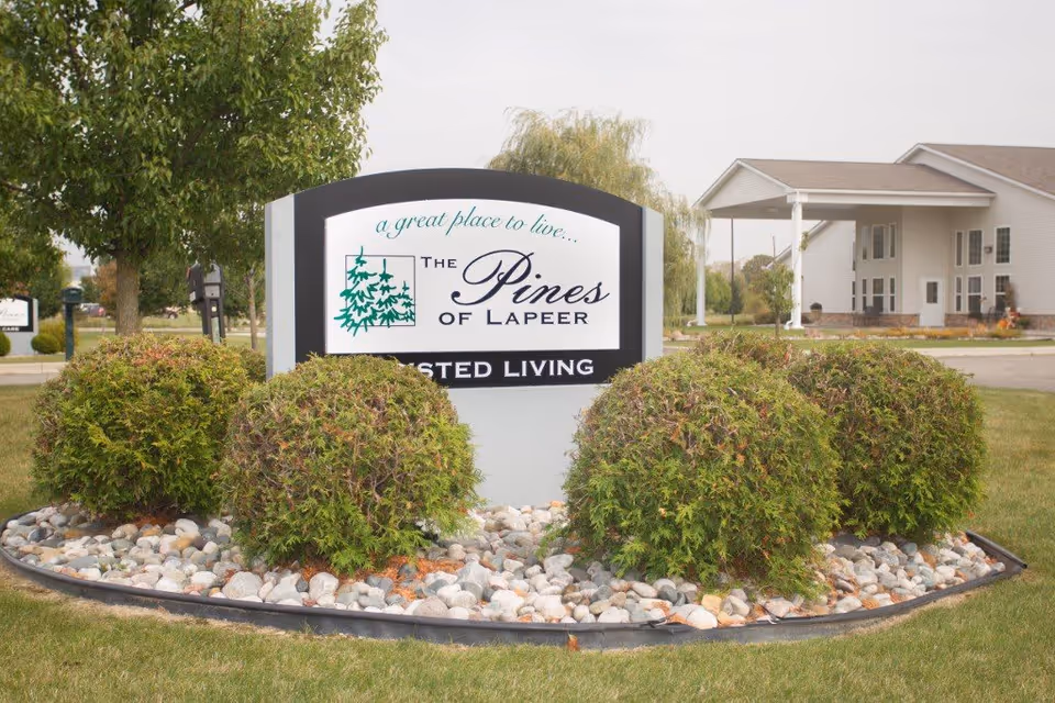 Entrance sign reading 'The Pines of Lapeer' for an assisted living facility set in a landscaped island with the building in the background.