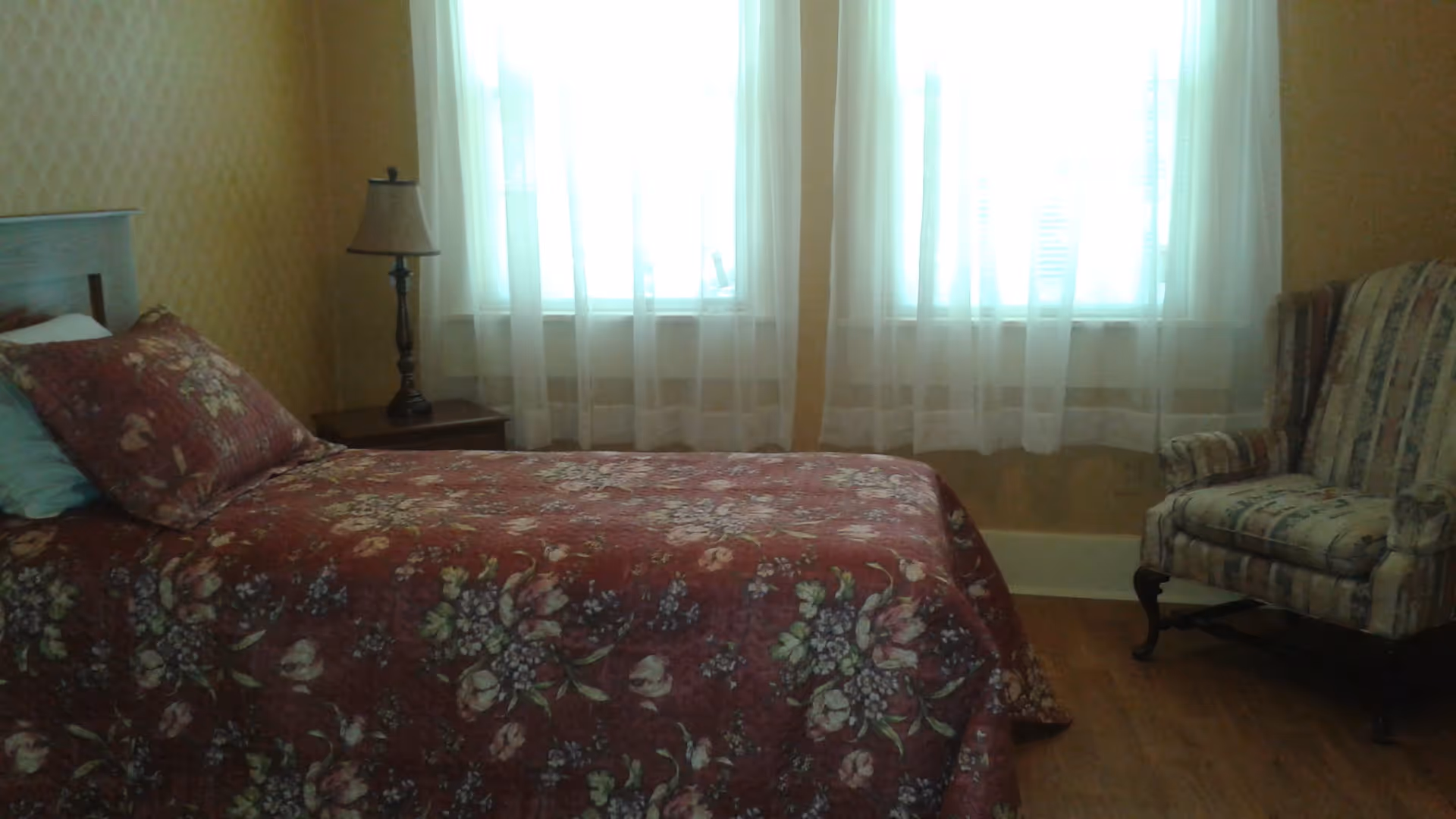 A bedroom with a floral-covered single bed, a bedside table and lamp, sheer-curtained windows, and an upholstered armchair.