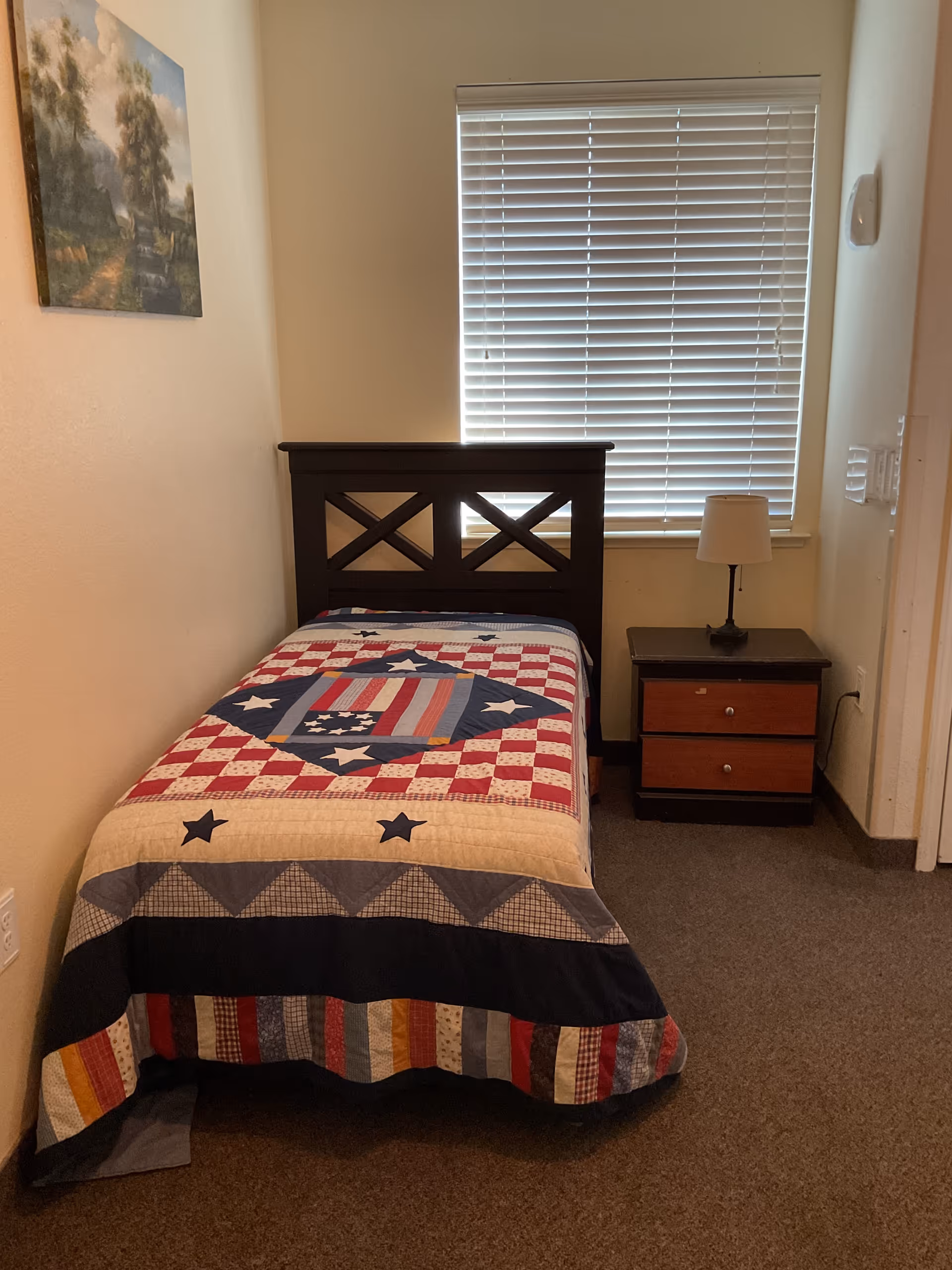 A small bedroom with a single bed covered by a patriotic quilt featuring stars and stripes. The bed has a dark wooden headboard with a crisscross design. Next to the bed is a wooden nightstand with two drawers and a small lamp on top. A window with closed blinds is behind the nightstand, and a landscape painting hangs on the wall to the left.