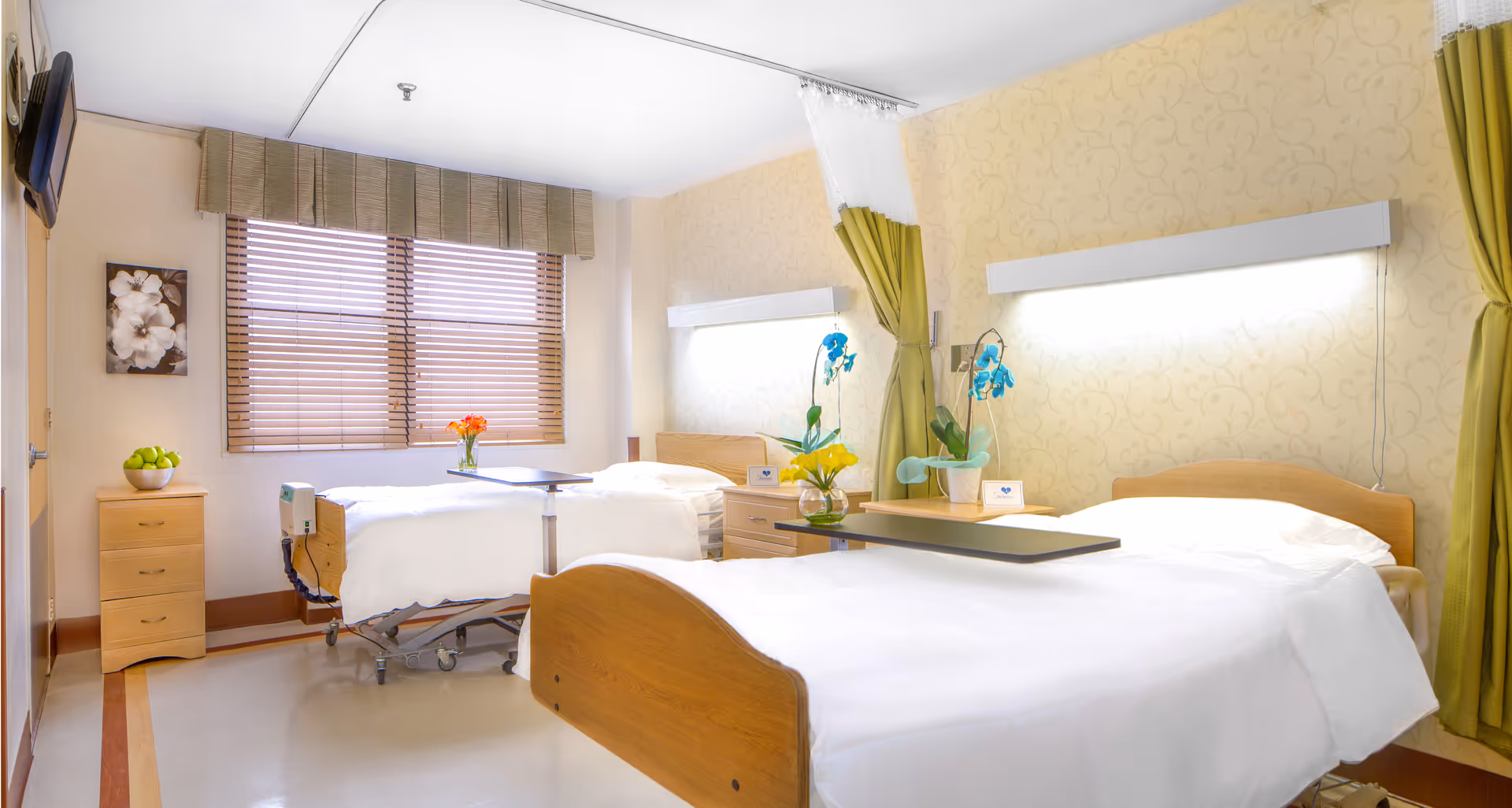 A bright and clean nursing home room with two hospital-style beds, each with white bedding and a small overbed table. There are bedside tables with flowers and medical equipment. The room has large windows with wooden blinds, light yellow patterned wallpaper, and green curtains. A small dresser with a bowl of green apples is also visible, along with a wall-mounted TV.