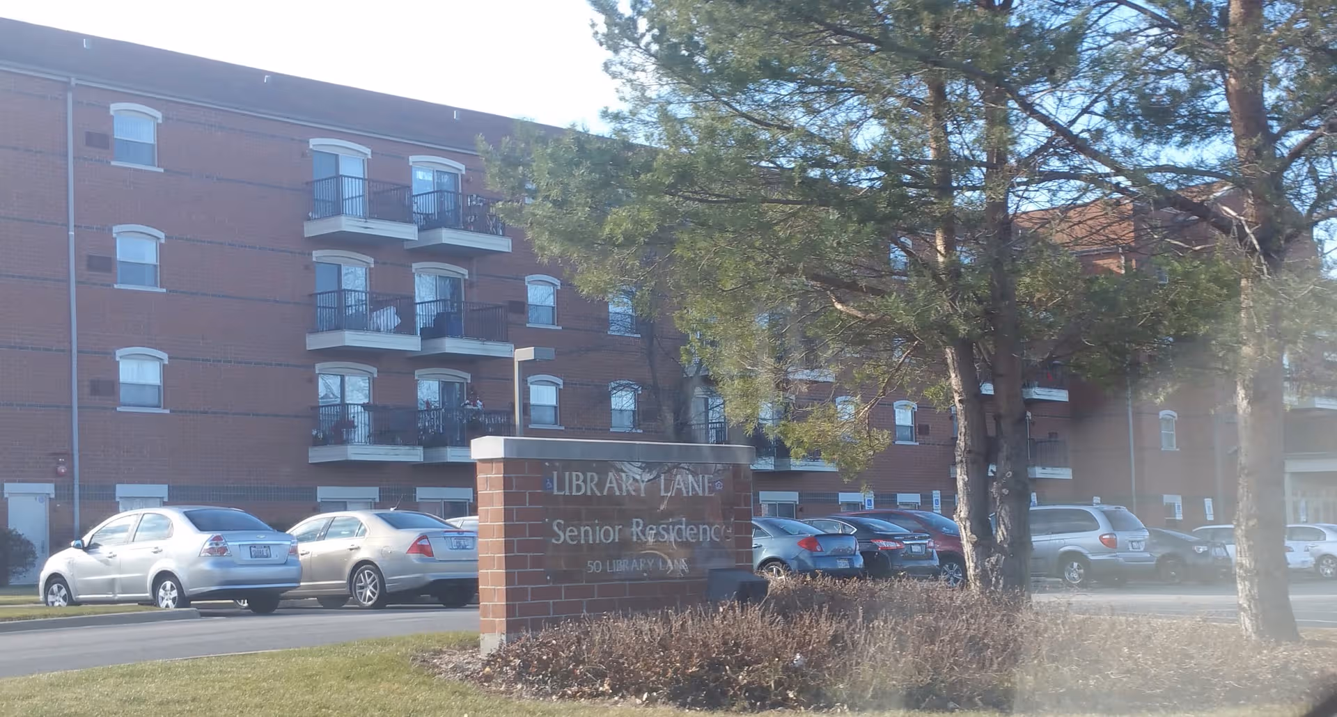 Brick senior residence building with balconies, a driveway, and a sign reading Library Lane Senior Residence.