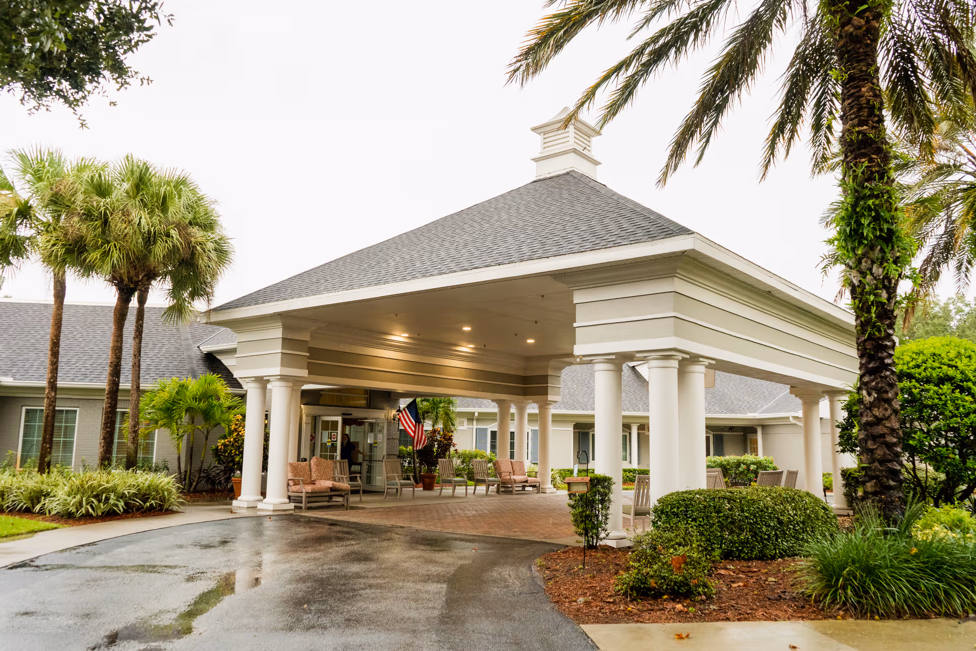 Covered entrance with white columns, palm trees, and seating leads into a senior living facility.