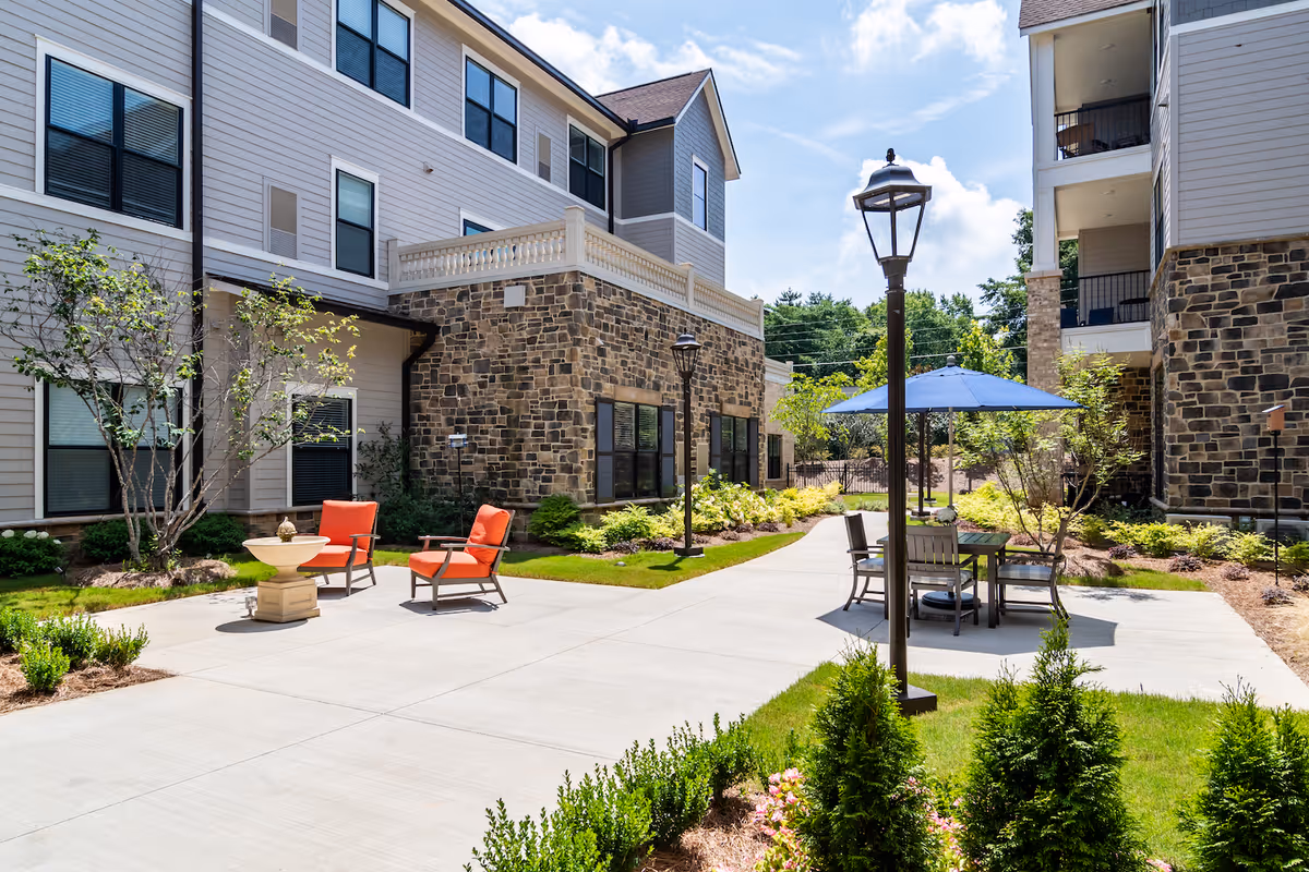 Outdoor courtyard area at The Solana East Cobb featuring a paved walkway, two orange cushioned chairs around a small fountain, a table with four chairs under a blue umbrella, surrounded by greenery and multi-story buildings with stone and siding exteriors.