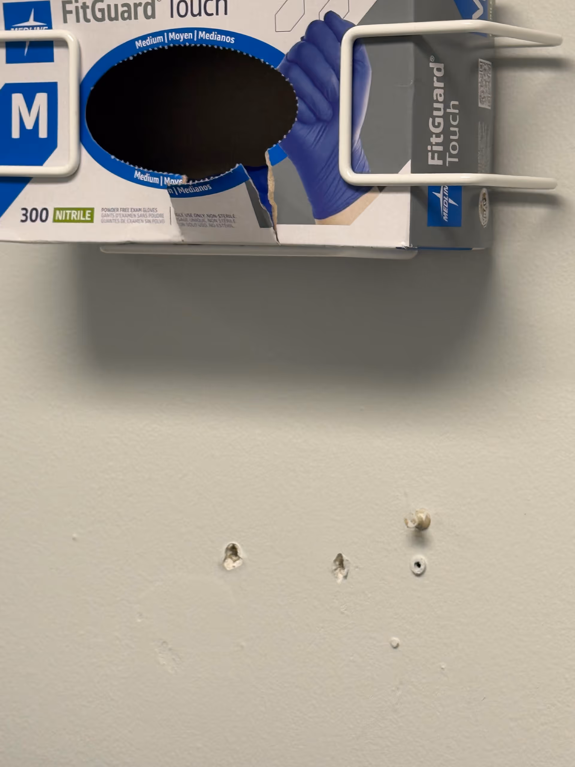 Wall-mounted box of nitrile gloves with torn packaging and damaged wall beneath.
