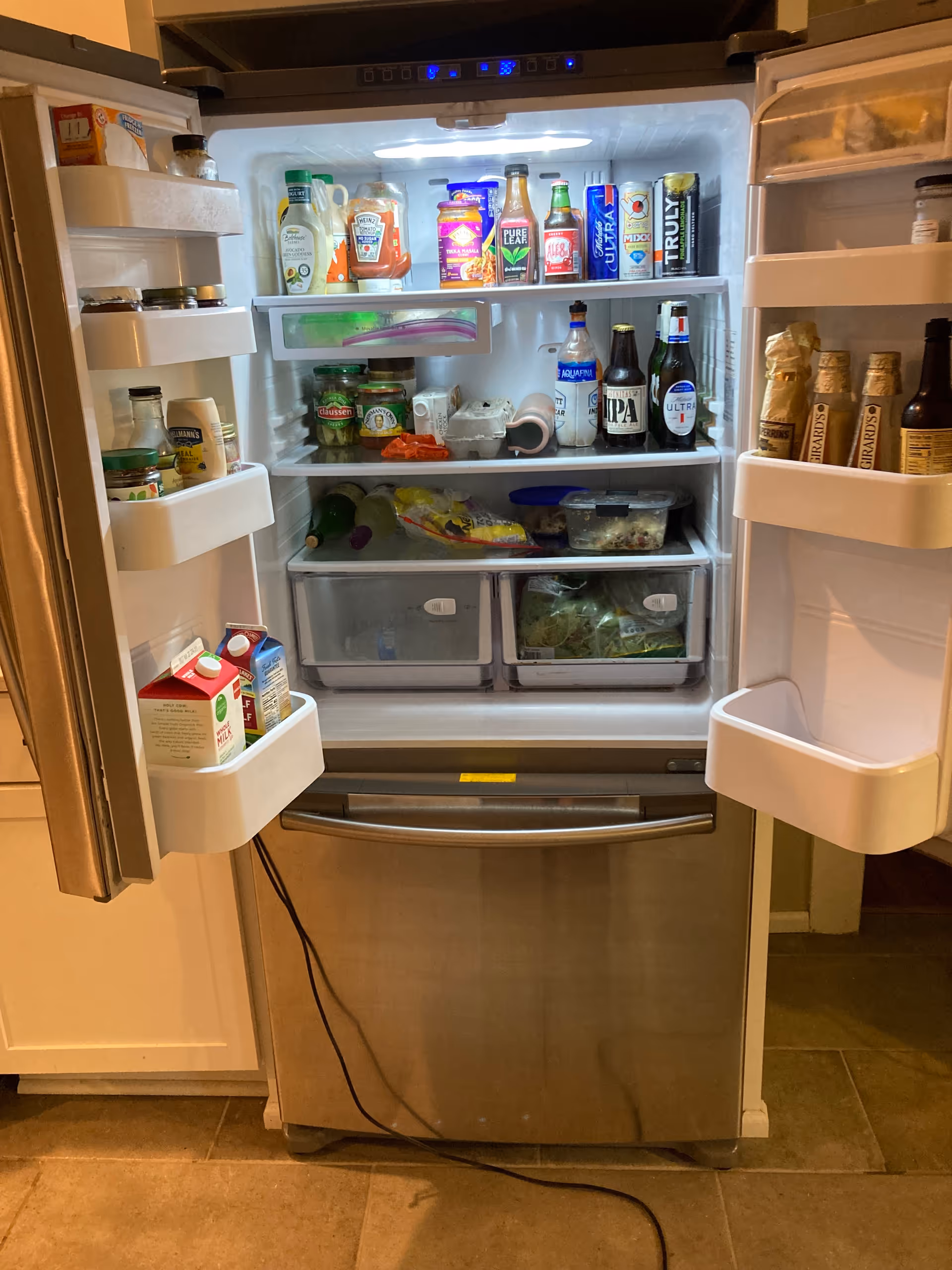 Open stainless steel refrigerator filled with condiments, drinks, and groceries in a kitchen.