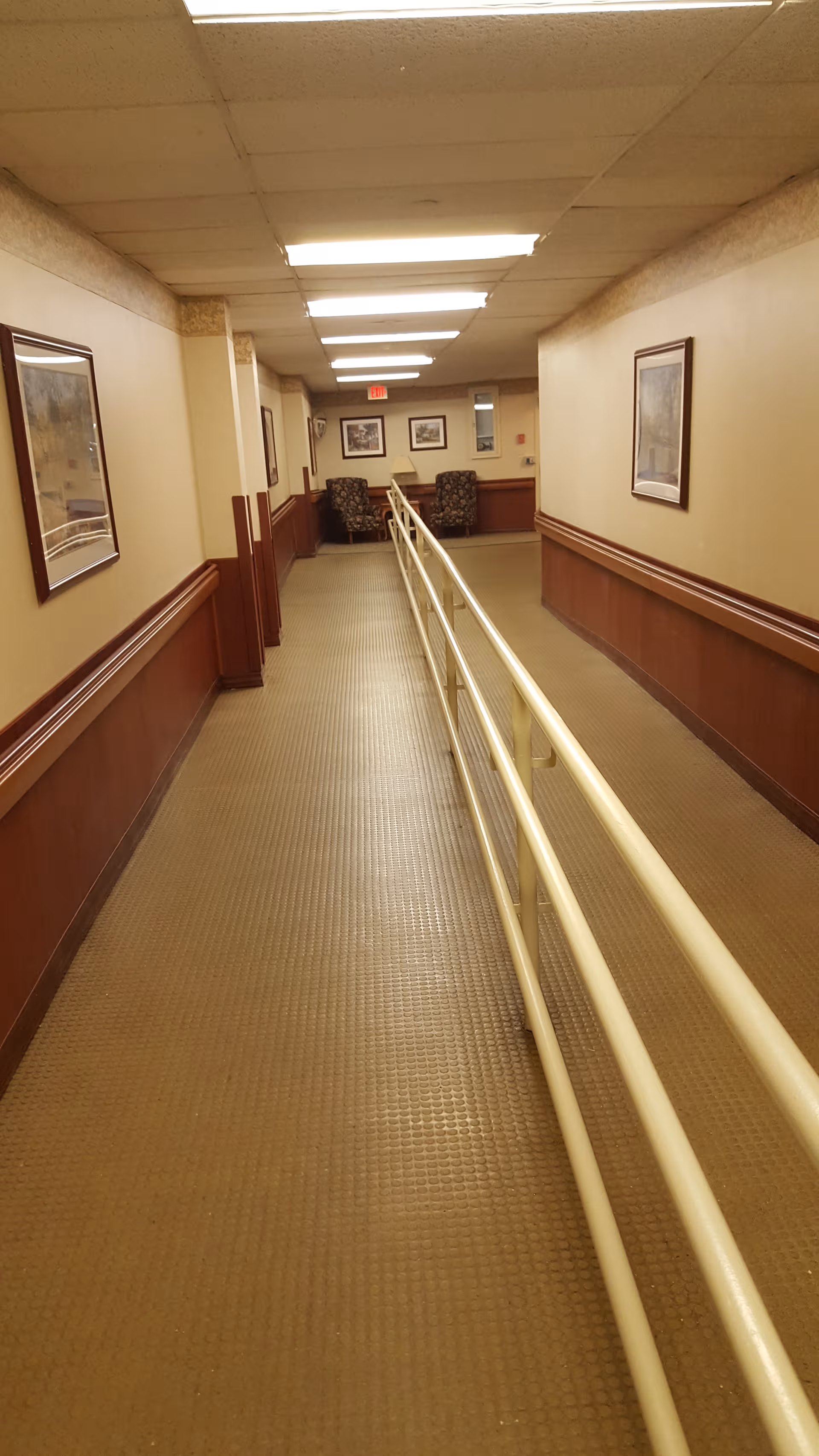 Indoor corridor of a senior facility with handrails, framed pictures, and distant seating.