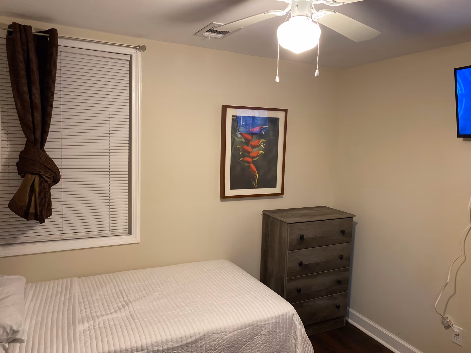 Cozy bedroom with a bed, dresser, window blinds, and ceiling fan in a neutral-colored room.