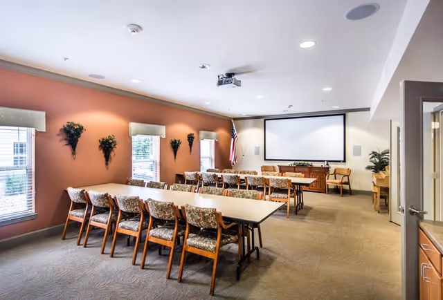 Large senior community meeting room with two long tables and floral-upholstered chairs facing a projection screen and an American flag.