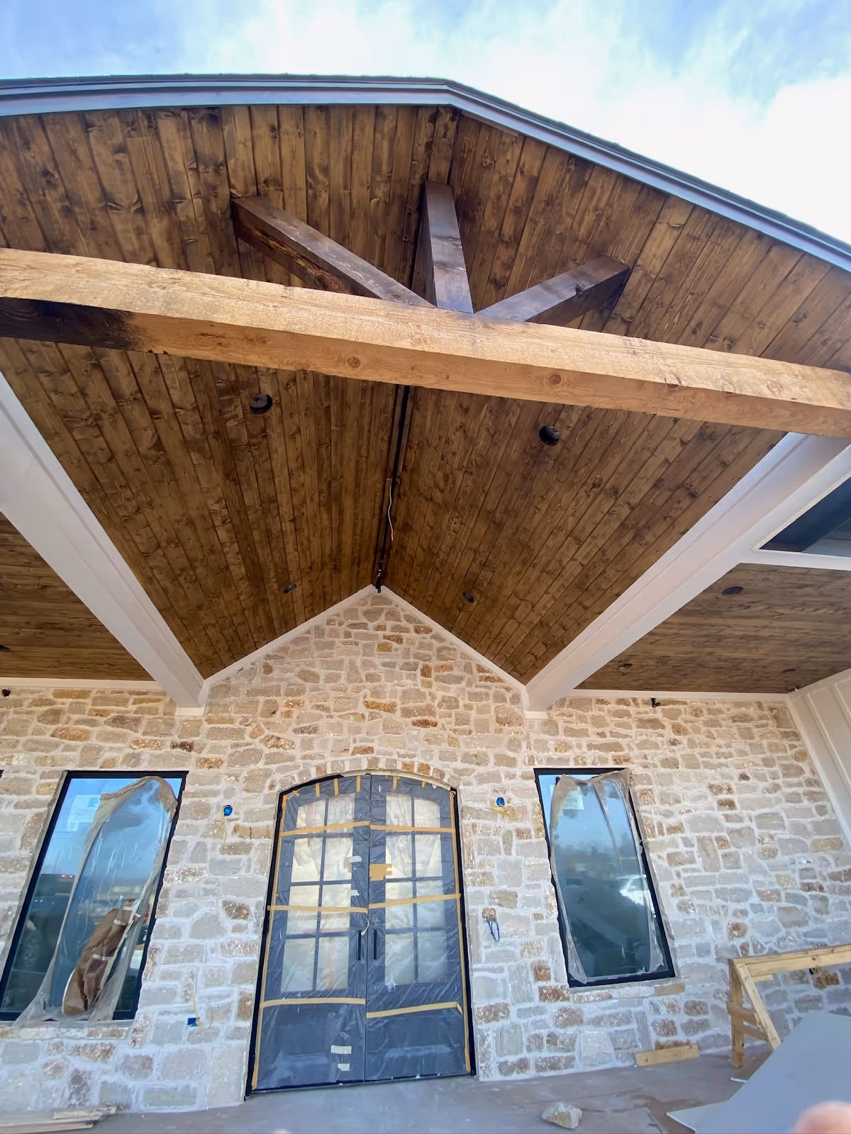Stone façade with a wooden peaked roof overhang and plastic-covered front doors under construction.