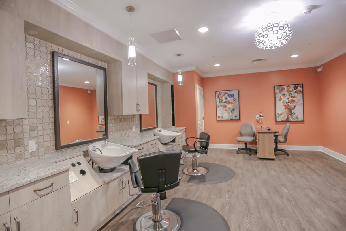 Interior view of a senior living facility's hair salon area with two black salon chairs in front of sinks and mirrors, light wood cabinetry, and a coral-colored wall with two floral paintings and a small desk with two gray chairs.