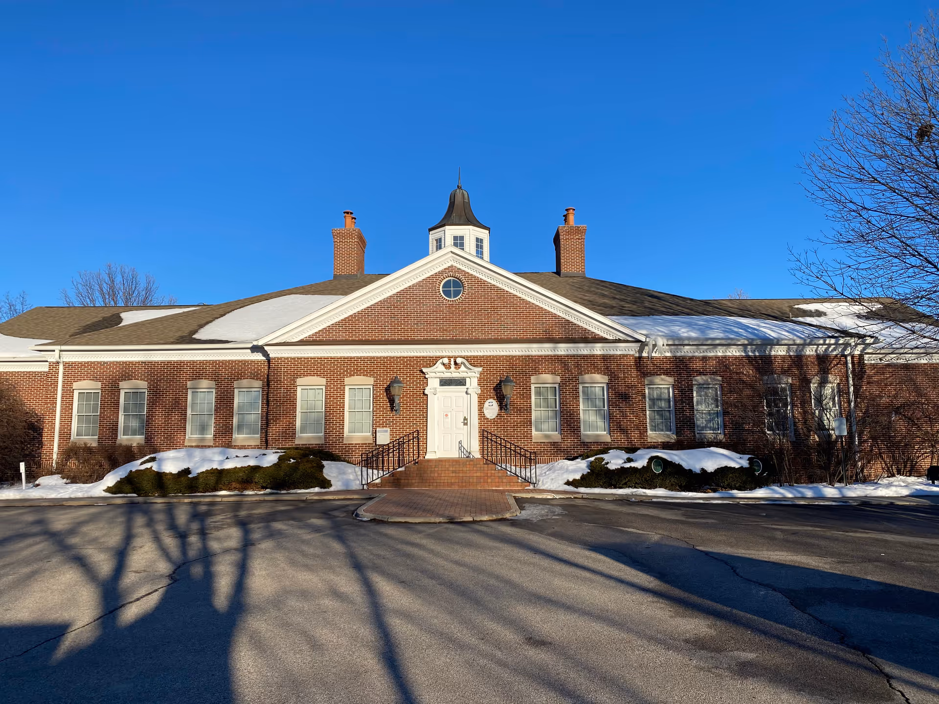 Brick senior living facility exterior with front entrance, steps, snow, and blue sky above.