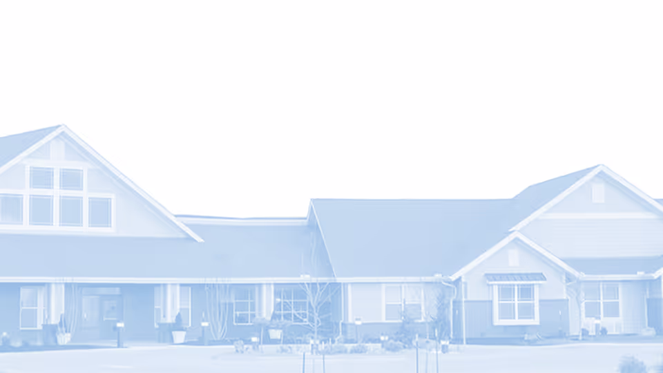 Exterior view of a single-story senior living facility building with multiple windows, pitched roofs, and a covered entrance area. The building is surrounded by small plants and shrubs.