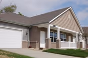 Exterior view of a single-story residential building with a brick facade and beige siding, featuring a white garage door, a white front door, and a small porch with white railings under a cloudy sky.