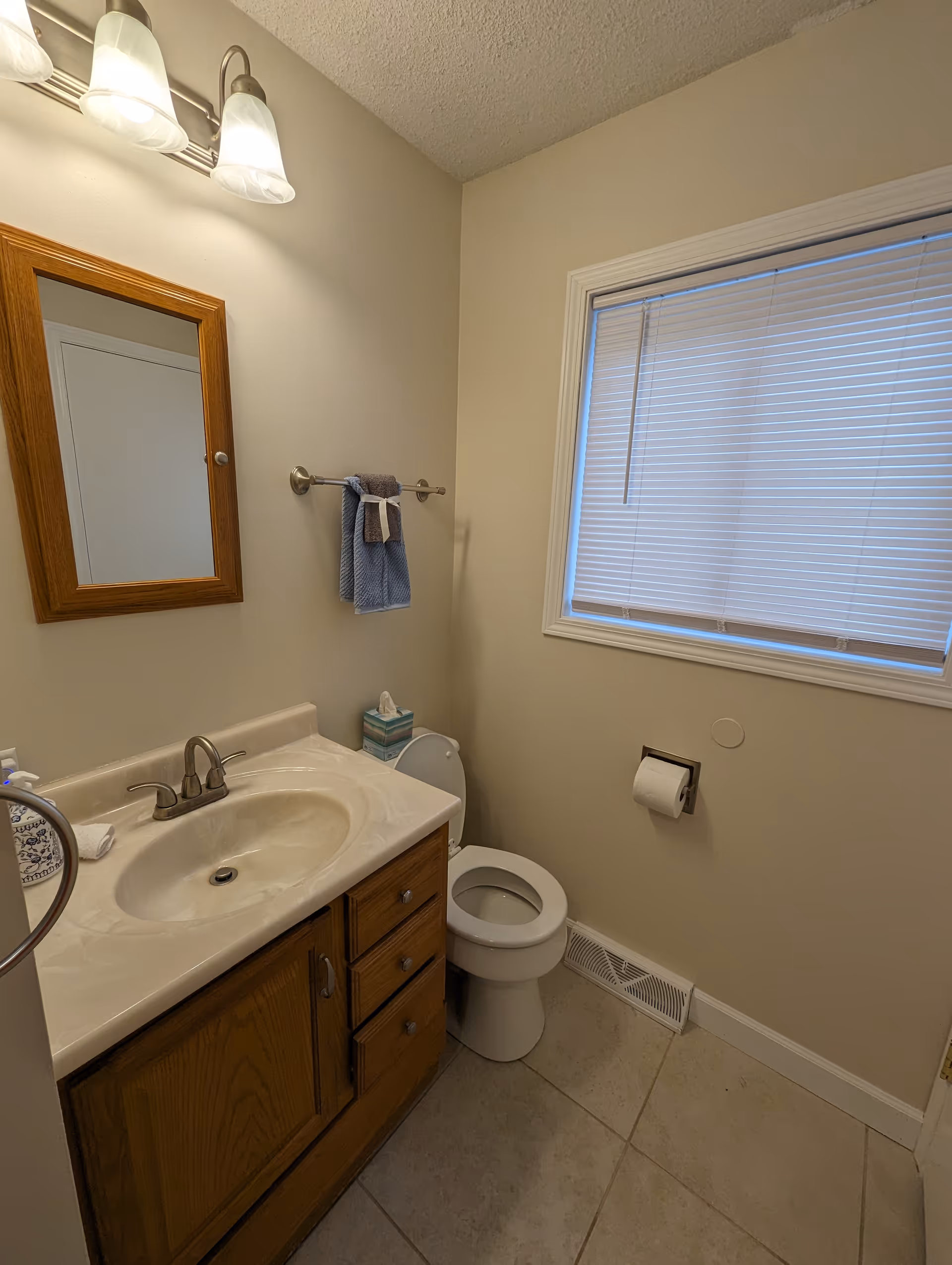 Small bathroom with sink, mirror, toilet, and window blinds in a senior living facility.