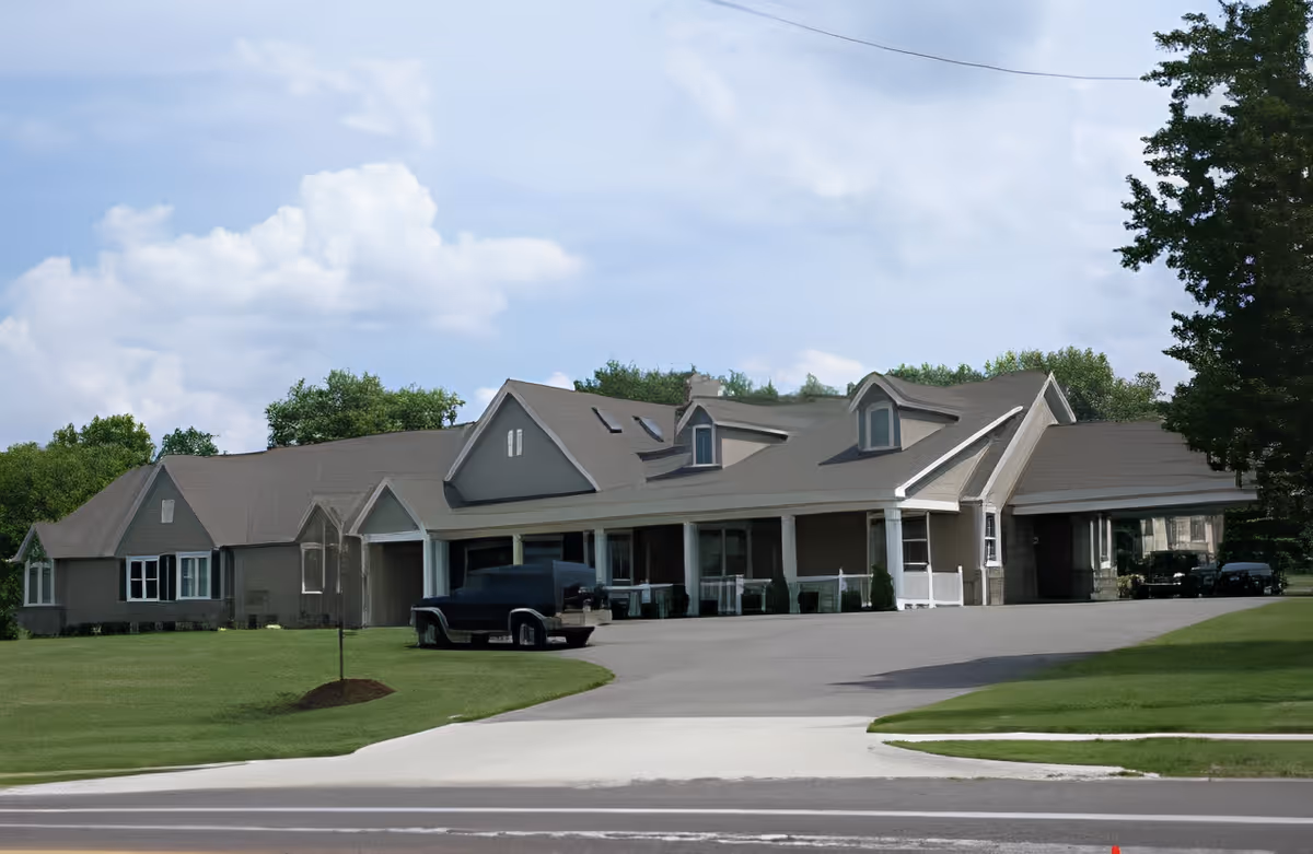Exterior view of a single-story building with a large driveway and a parked black vehicle in front. The building has a gray roof with dormer windows and a covered porch area. There is green grass and trees surrounding the building under a partly cloudy sky.