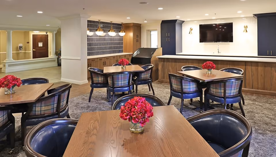 A cozy dining area with wooden tables and plaid cushioned chairs arranged on a carpeted floor. Each table has a small vase with bright pink flowers. The background features a counter with dark blue cabinets and a mounted flat-screen TV above it, along with pendant lights and a tiled backsplash on the left side.