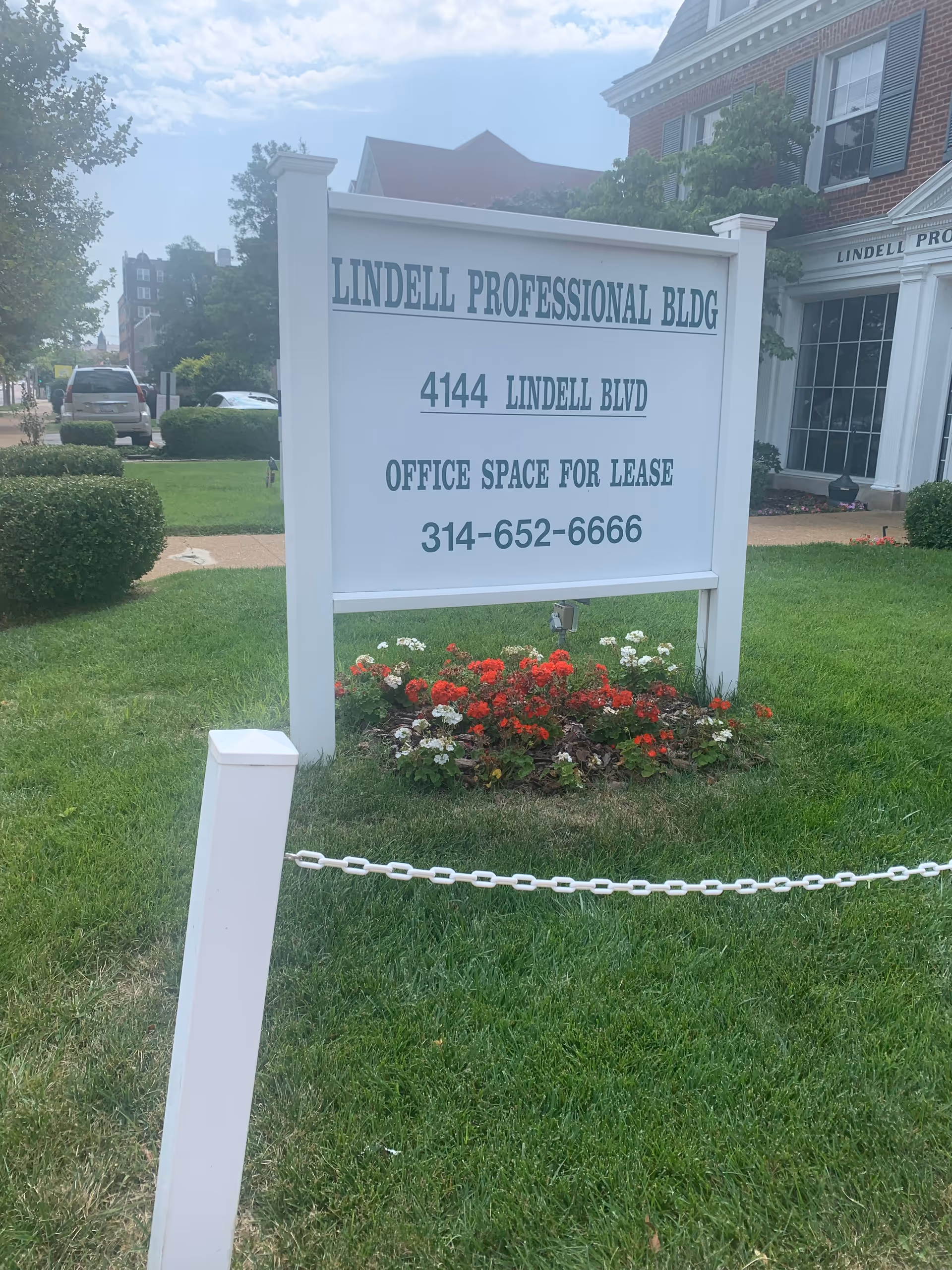 Exterior sign and flower bed in front of a brick building with greenery.