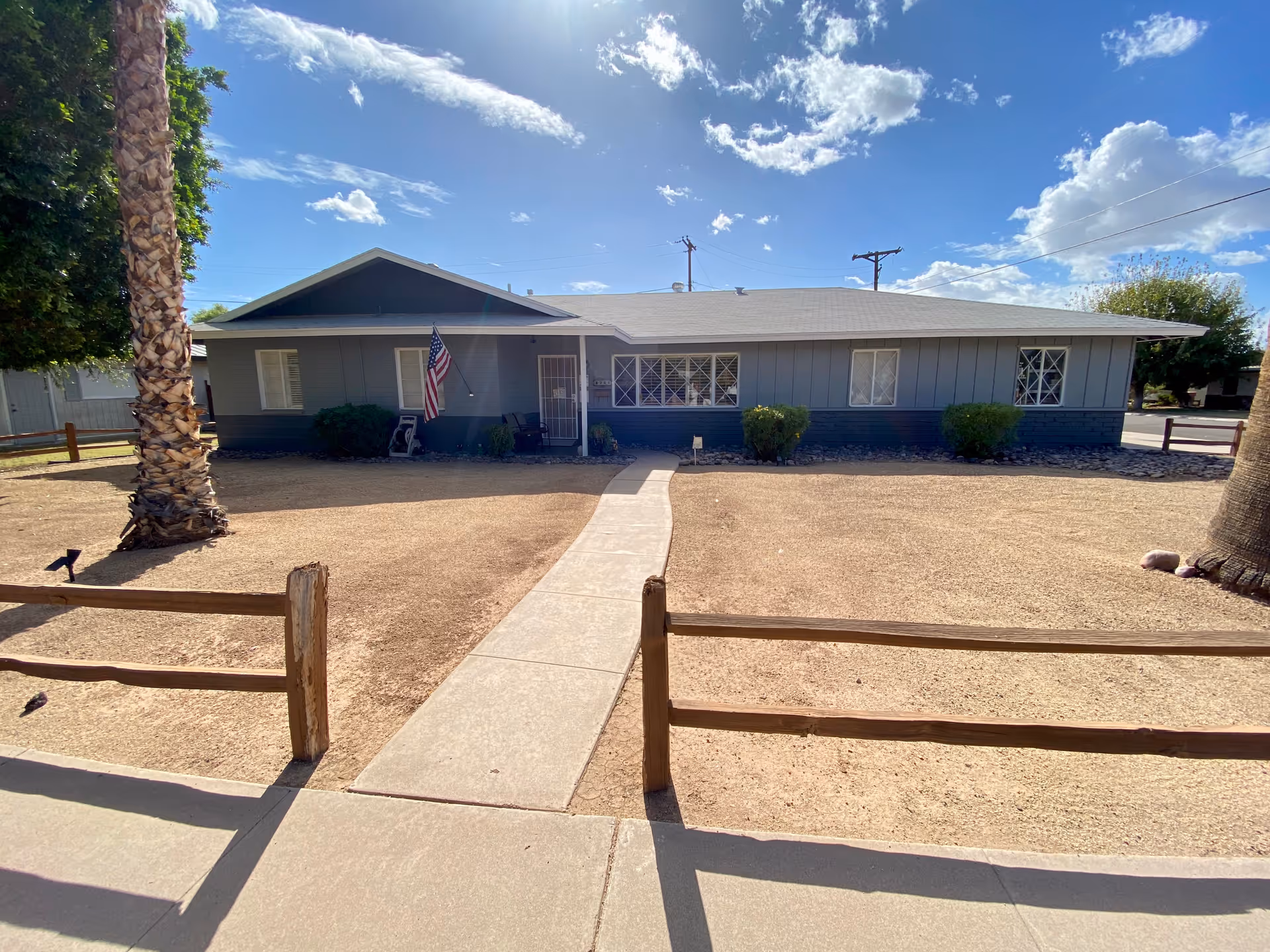 Single-story building with a gray exterior and a white roof under a bright blue sky with scattered clouds. A concrete walkway leads from the sidewalk to the front door, which has an American flag hanging nearby. The yard is mostly bare dirt with a few bushes and two palm trees, and a simple wooden fence borders the property.