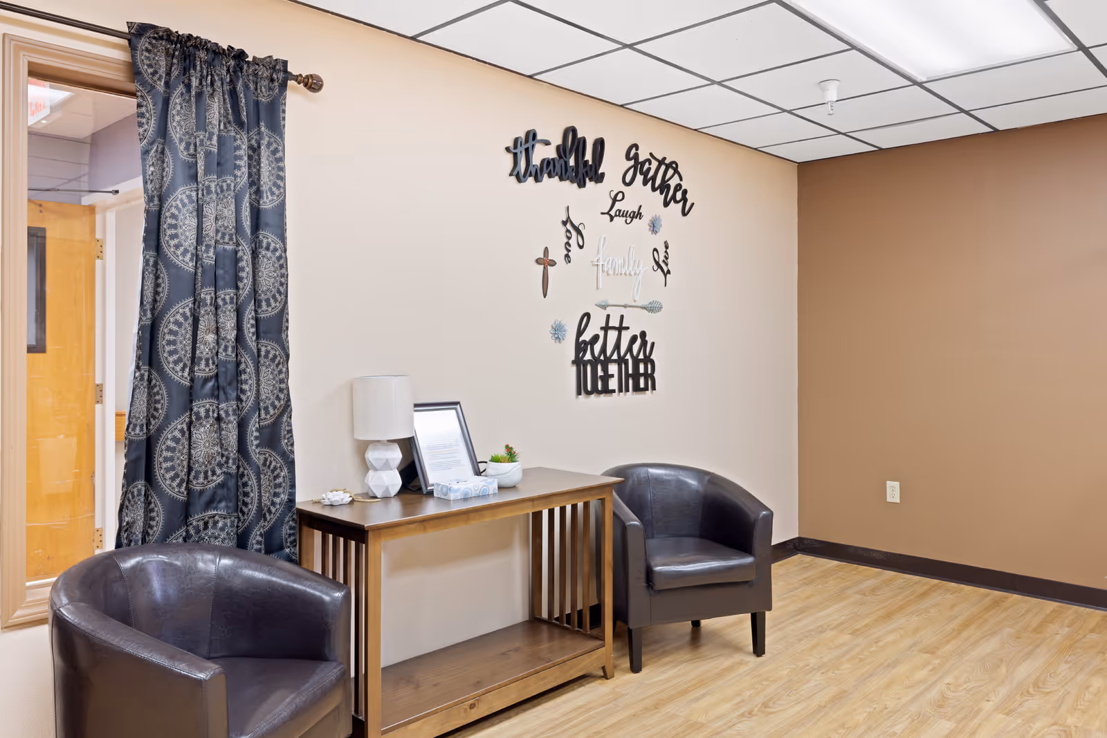 A small seating area in a room with two dark leather armchairs, a wooden console table with a white lamp, framed picture, and small plant. The wall behind the table has decorative words including 'thankful', 'gather', 'laugh', 'love', 'family', and 'better together'. There is a window with patterned dark curtains on the left side and light wood flooring.